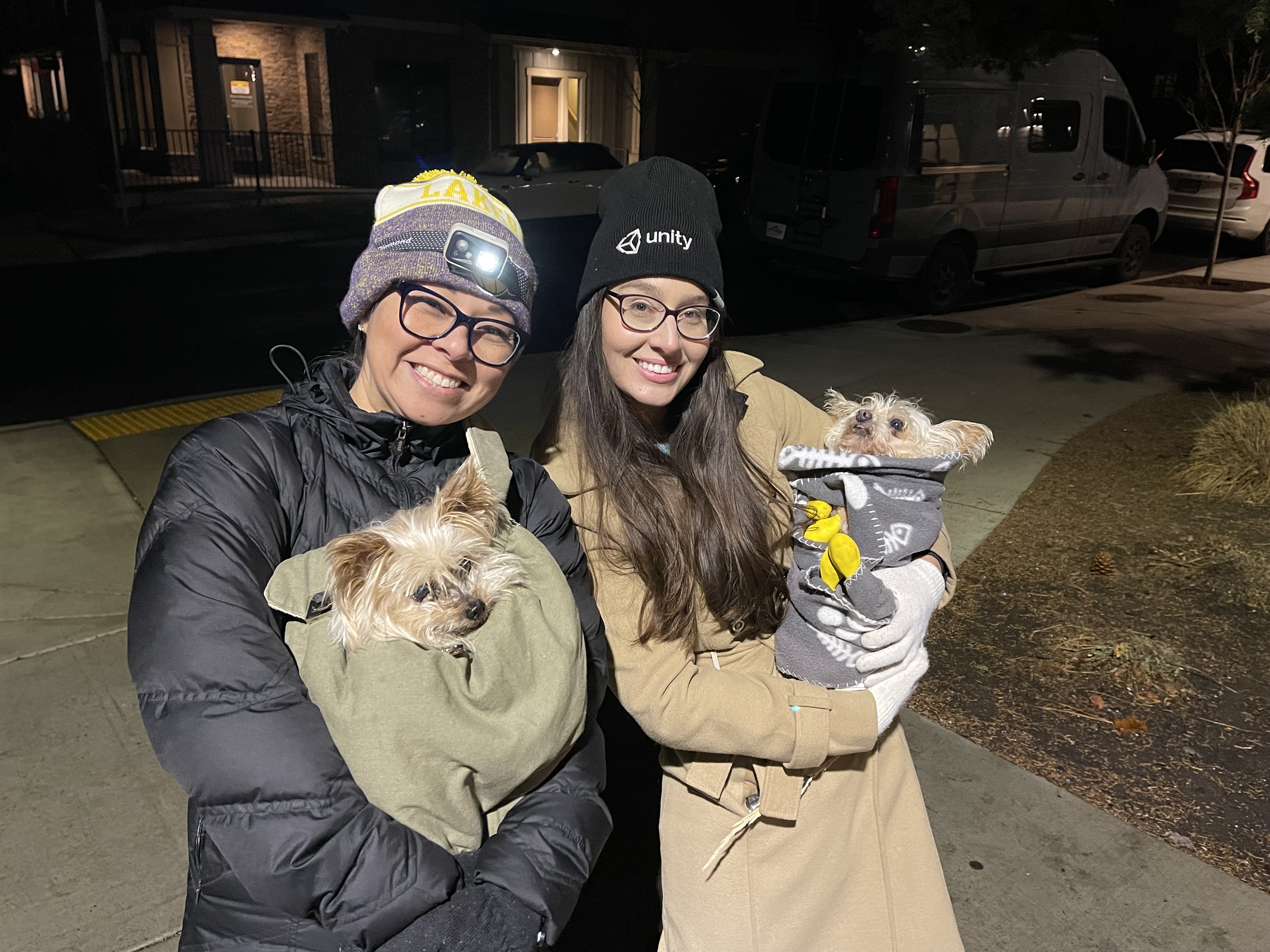 Two friends bundled up at night holding small dogs, Samantha wearing a Unity beanie